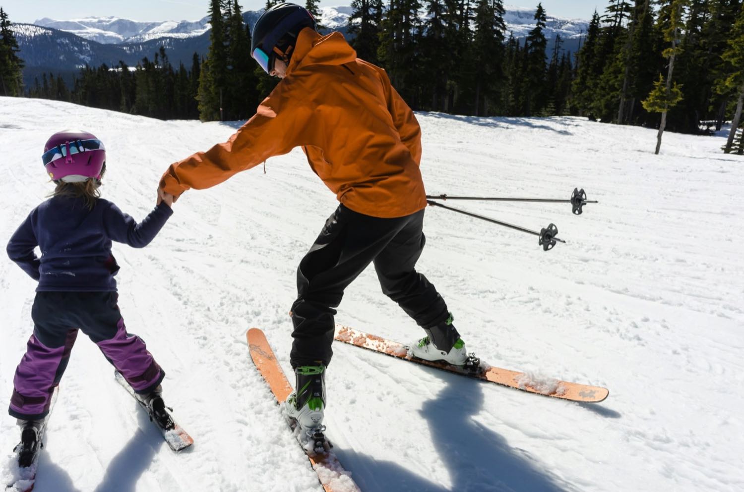 An adult in an orange jacket holding a small child's hand as she learns to ski on a gentle slope