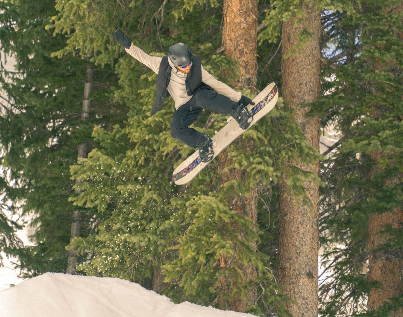 A snowboarder airborne off a jump with tall spruce and pine trees filling the background